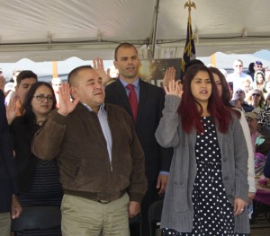 Hands raised to become the newest US citizens. Raul  Campos in brown jacket, Fabiano Menezes de Souza wearing a red tie, and Juliana Vaca-Triperri in the black and white dress.