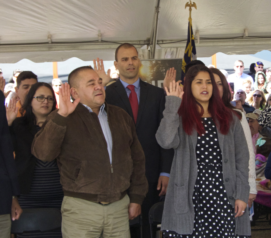Hands raised to become the newest US citizens. Raul Campos in brown jacket, Fabiano Menezes de Souza wearing a red tie, and Juliana Vaca-Triperri in the black and white dress.